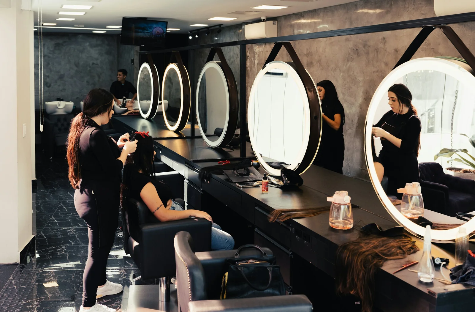 Stylist works with a client in a charcoal-toned salon lined with round mirrors